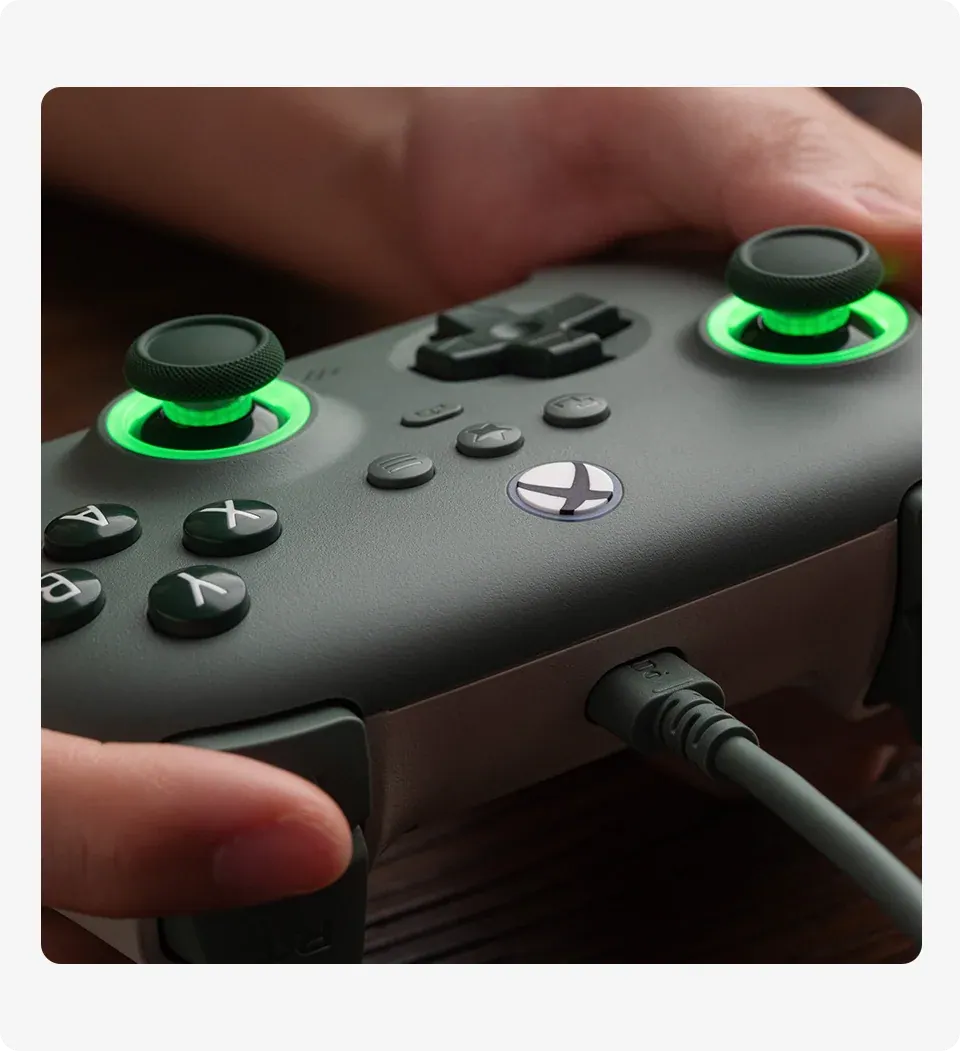 A top-down view of the 8BitDo Ultimate C Wired Dark Green controller on a wooden desk alongside a laptop and other controllers.