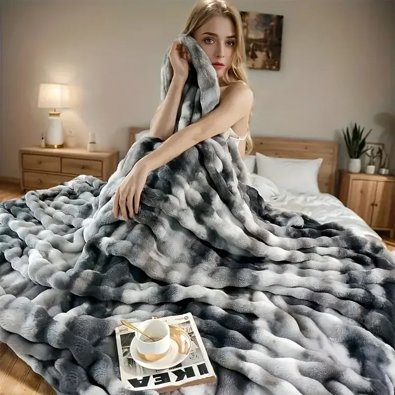 A woman wrapped in a cozy grey and white faux rabbit fleece blanket in a bedroom setting.