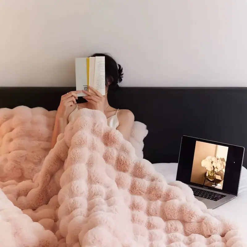 A girl sitting in bed covered with a soft light pink faux fleece blanket while reading.