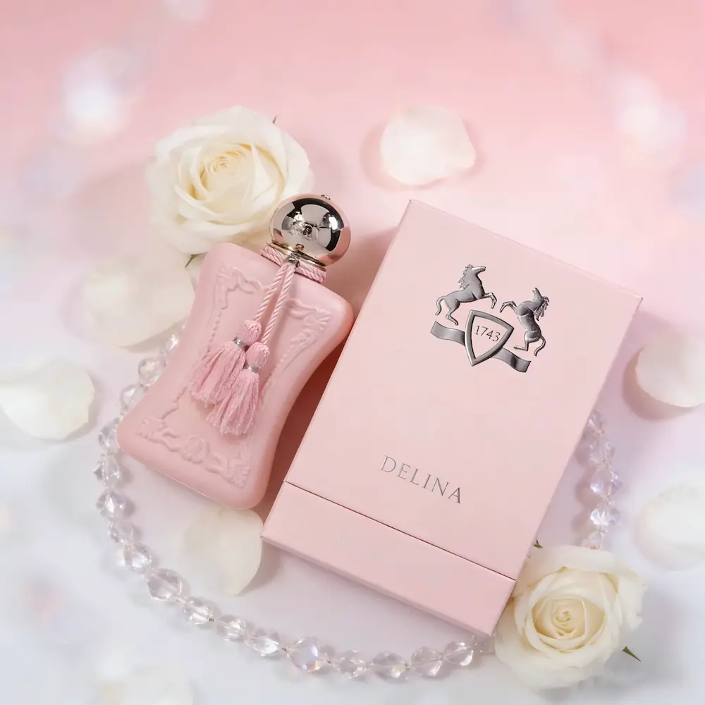 A flatlay composition of the elegant pink Delina perfume bottle, branded box, white roses, and a crystal necklace.